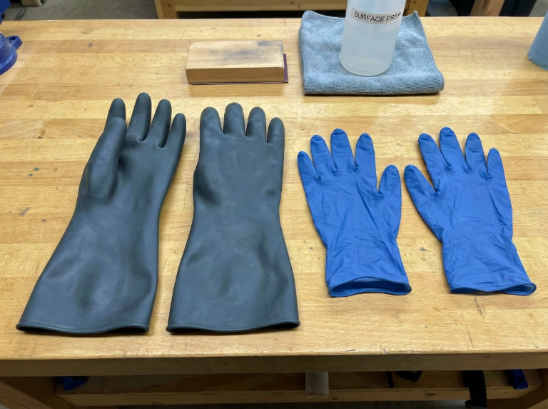 Bir çift uzun siyah bütil eldiven ve bir çift mavi nitril eldiven, bir mikrofiber bez ve yüzey hazırlama şişesinin yanında bir çalışma tezgahının üzerine serilmiş.